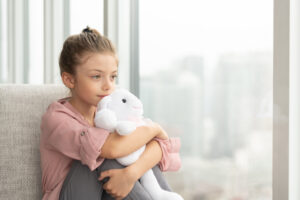 A cute elementary age girl is sitting near a large window looking at the urban skyline outside. Her arms are wrapped around her legs and she is holding a stuffed toy. She has a calm and reflective expression on her face.