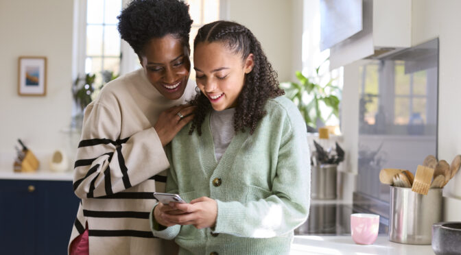 Mother Talking With Teenage Daughter At Home Looking At Social Media On Mobile Phone Together