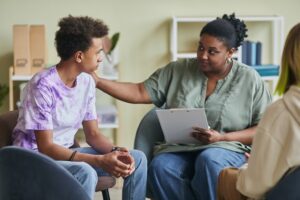 African American young psychologist talking to teenage boy during therapy session at classroom